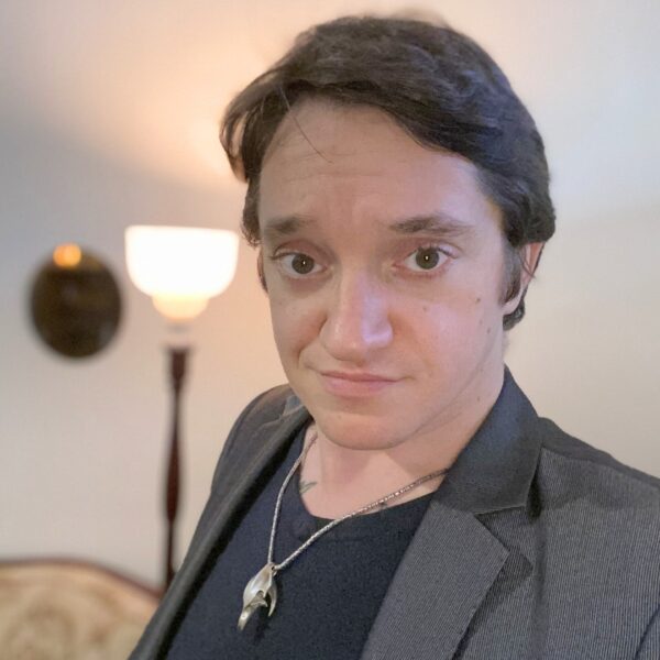 Photo of Matt, a young man with dark brown hair wearing a grey suit jacket and black top underneath. He is looking directly at the camera with a slight smile. There is a lit lamp in the background.