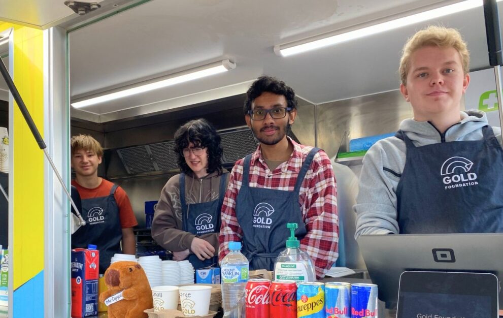 Four young people wearing Gold Foundation aprons stand inside a food van, smiling at the service window. Cups, drink cans and café supplies sit on the counter in front of them.