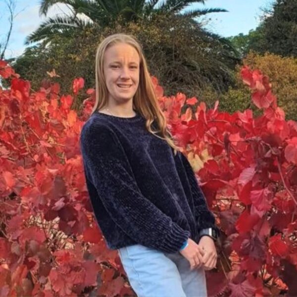 A young woman with long blonde hair stands outdoors smiling in front of vibrant red autumn foliage, wearing a dark navy knit jumper and jeans, with trees and greenery behind her.