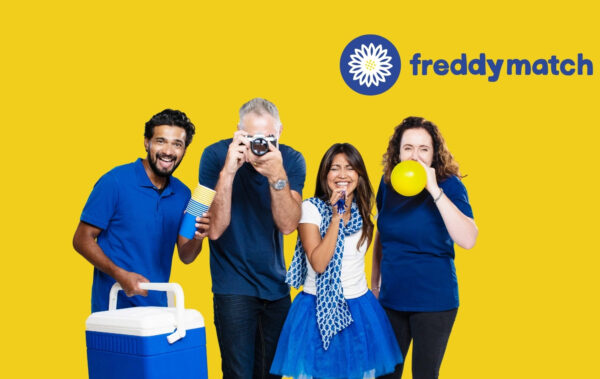 Four people dressed in blue and white stand cheerfully in front of a bright yellow background. They are holding party items including a cooler, cups, a camera, a pom-pom, and a yellow balloon. All are smiling or laughing. In the top left corner, the logo and name 
