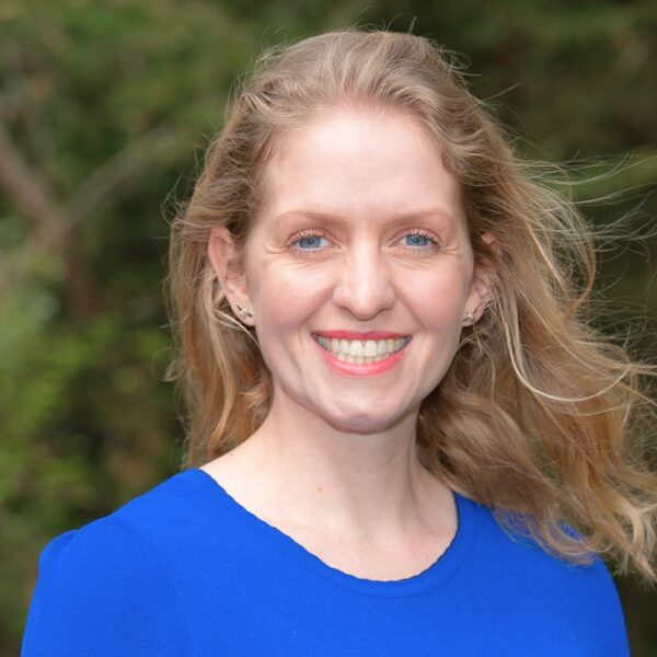 A woman with long, wavy blonde hair smiles brightly while standing outdoors. She is wearing a vibrant blue top and small earrings. The background is soft-focus greenery.