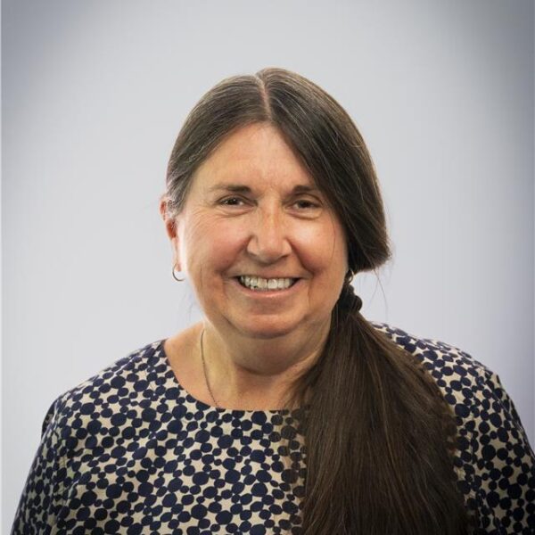 Portrait of a smiling woman with long dark hair over one shoulder, wearing a patterned top against a plain background.