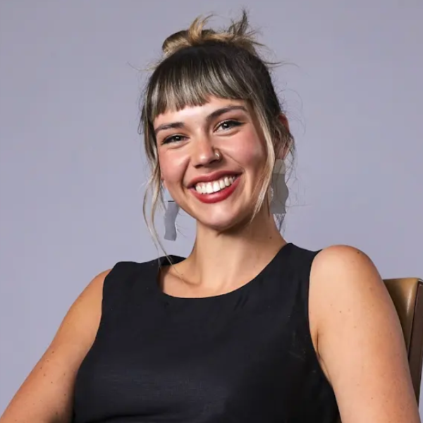 Portrait of a smiling femme presenting person with dark hair tied in a bun and straight-cut fringe, wearing a sleeveless black top, long statement earrings and a nose ring against a soft grey background.